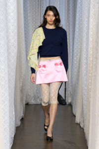 Model walks the runway in a navy cropped top, pink satin mini skirt with bow pockets, lace leggings, and a yellow printed one-shoulder cape.