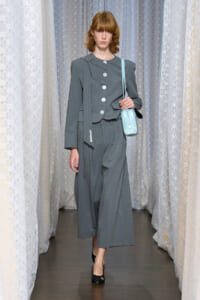 Model walking a runway in a gray wide-leg suit with an oversized blazer, carrying a light blue small shoulder bag.