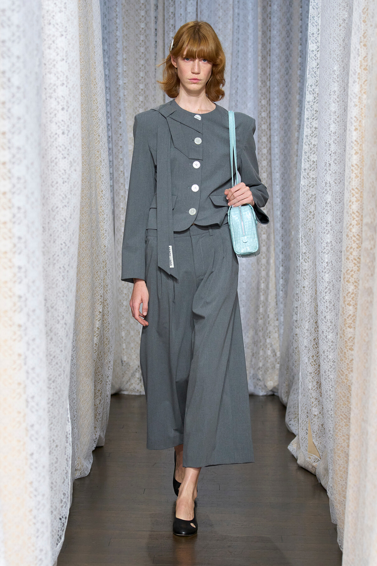 Model walking a runway in a gray wide-leg suit with an oversized blazer, carrying a light blue small shoulder bag.