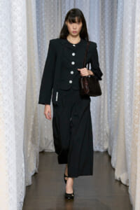 Female model walking a runway in a black tailored suit with oversized silver buttons, carrying a dark brown textured handbag.