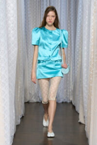 Runway model in a turquoise satin two-piece set with puff sleeves, lace tights, white flats, and a small round purse.