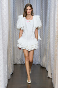 Model walking runway in a white Victorian-inspired dress with oversized puff sleeves and tiered ruffled skirt.