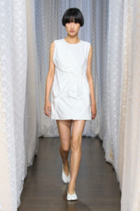 Model walking a runway in a sleeveless white mini dress with sculpted draping on the front, paired with white flats.