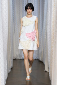Model walking a runway in a white sleeveless dress with cream lace appliqués and a pink gingham detail on the front, in a studio setting with lace curtains.