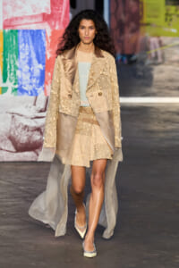 Model walking a fashion show in a beige lace blazer with satin lapels and a sheer, flowing cape over a pale dress, against a colorful backdrop.