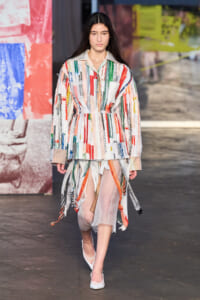 Female model walking a fashion runway in a multicolored striped jacket with a sheer, layered skirt and white heels.