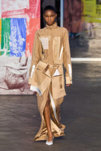 Female model walking a runway in a beige patchwork outfit with asymmetrical wrap skirt, white patches, and long sleeves, paired with white heels.