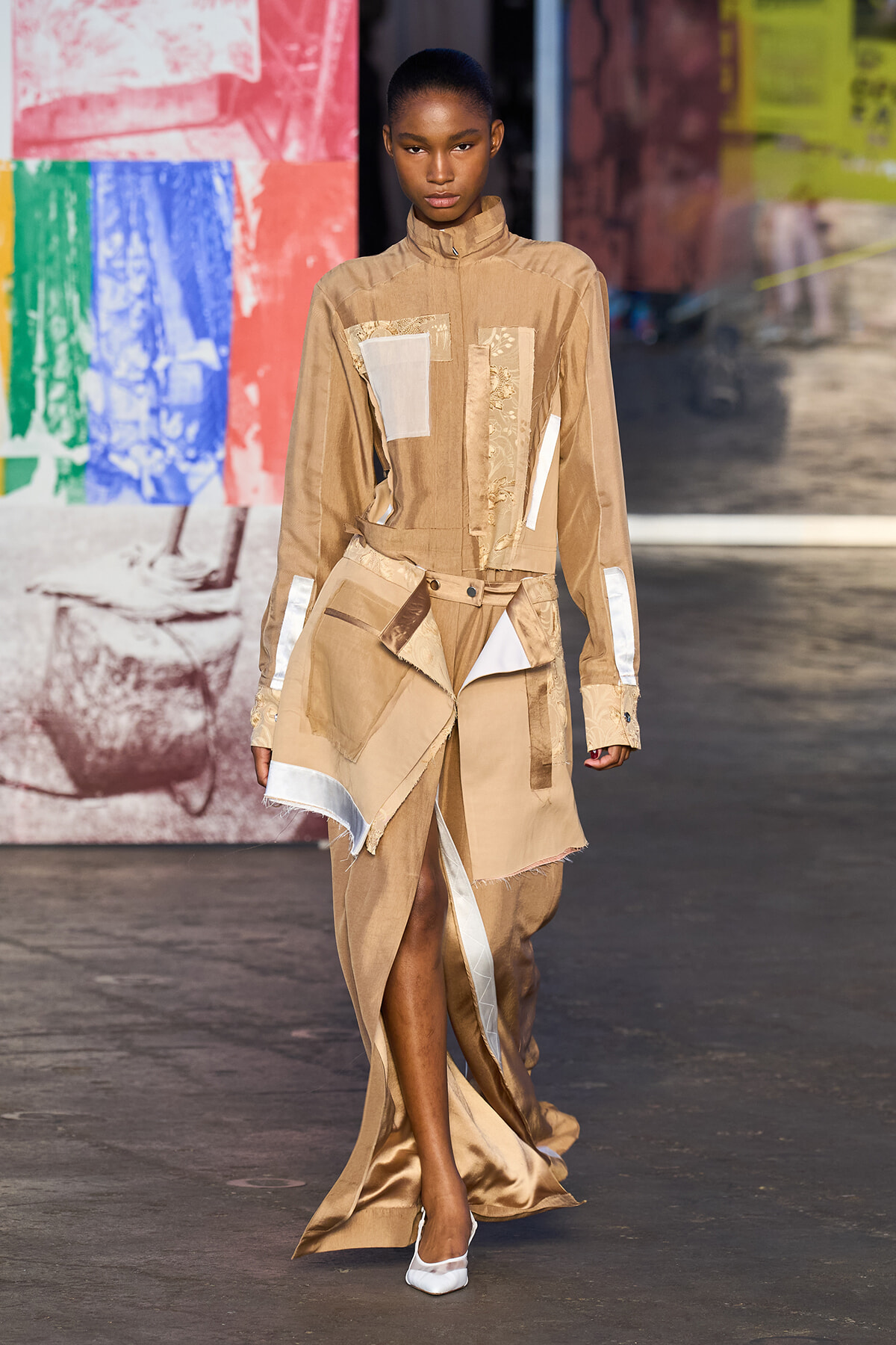 Female model walking a runway in a beige patchwork outfit with asymmetrical wrap skirt, white patches, and long sleeves, paired with white heels.