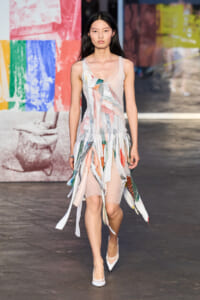 Model walking runway in a sheer, multi-colored fringe dress with white heels; colorful wall backdrop behind her.