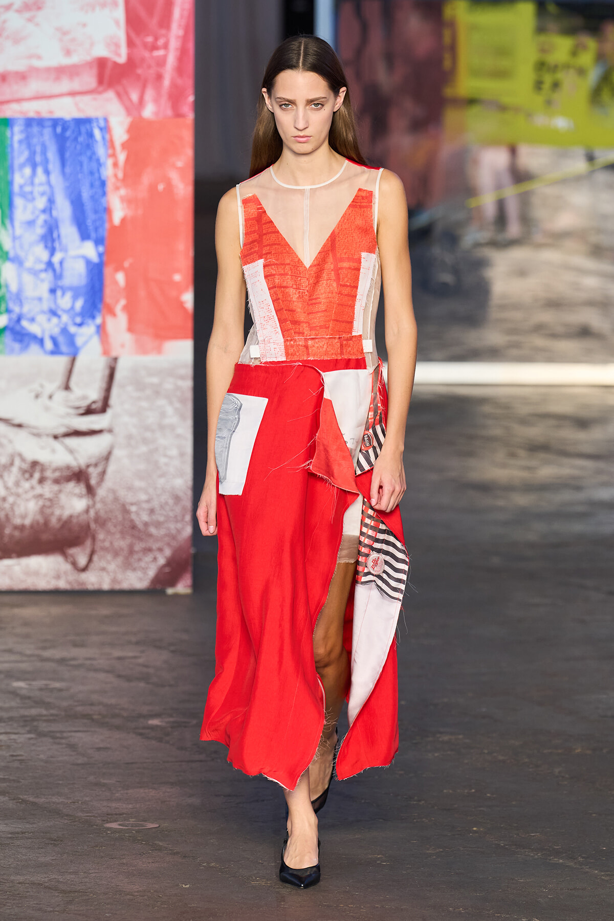 Model walking a runway in a red patchwork maxi dress with sheer panels and a V-neckline at a fashion show.