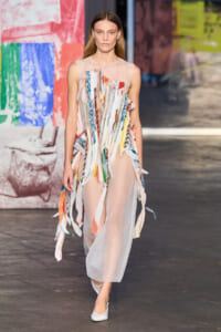 Model walking the runway in a sheer pale-pink dress covered with multicolored fabric ribbons.