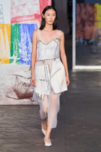 Model walks the runway in a white layered outfit with sheer panels, against a colorful art backdrop.