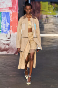 Model walks a runway in a beige trench coat with color-block pockets over a lace two-piece set, paired with pale heels.