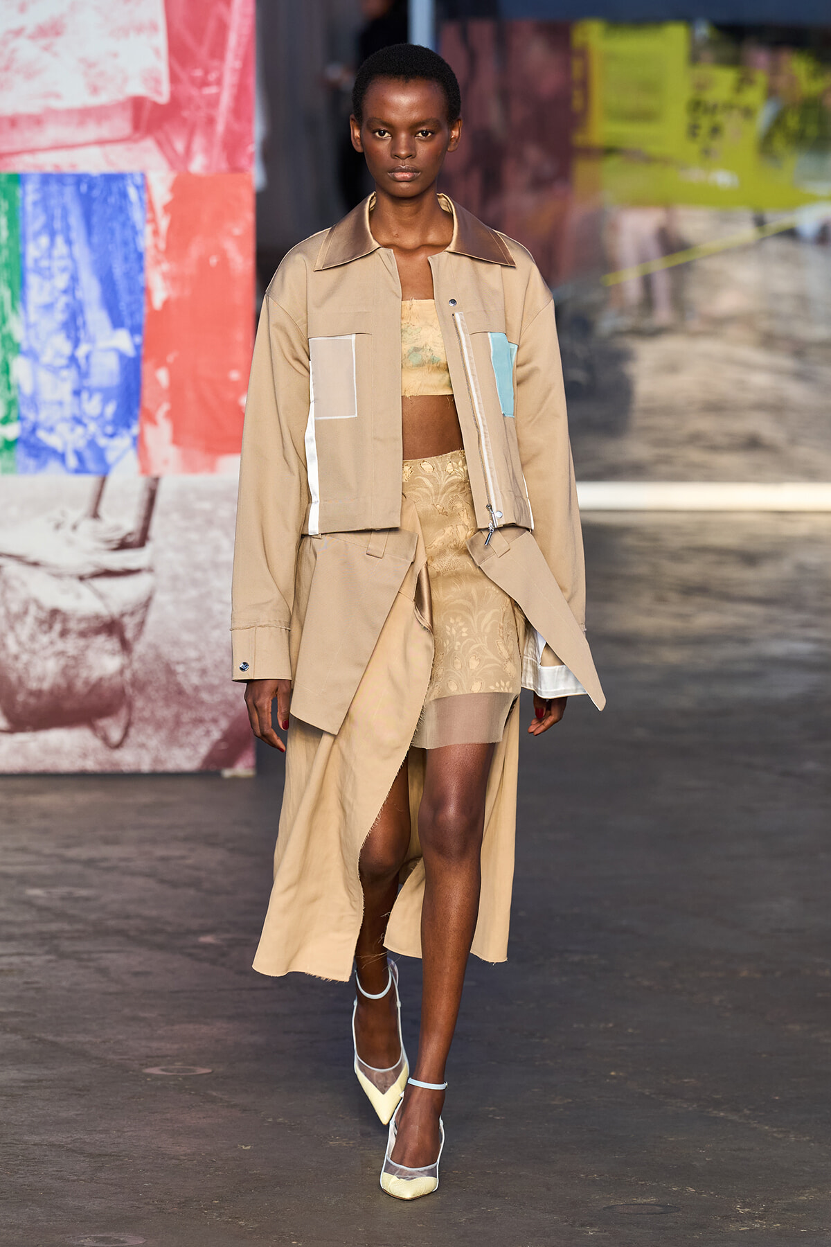 Model walks a runway in a beige trench coat with color-block pockets over a lace two-piece set, paired with pale heels.