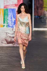 Model walking a runway in a pastel patchwork dress and pale blue bralette against a colorful mural backdrop