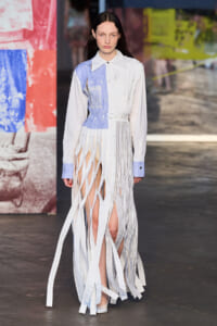 Model walks a fashion show wearing a mixed-white and light-blue shirt-dress with cut-out and ribbon-like fabric strips cascading to the floor, creating a fringe effect.