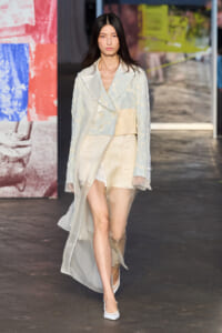 Female model walking a fashion runway in a pale blue-beige layered ensemble with a sheer cape and white heels.
