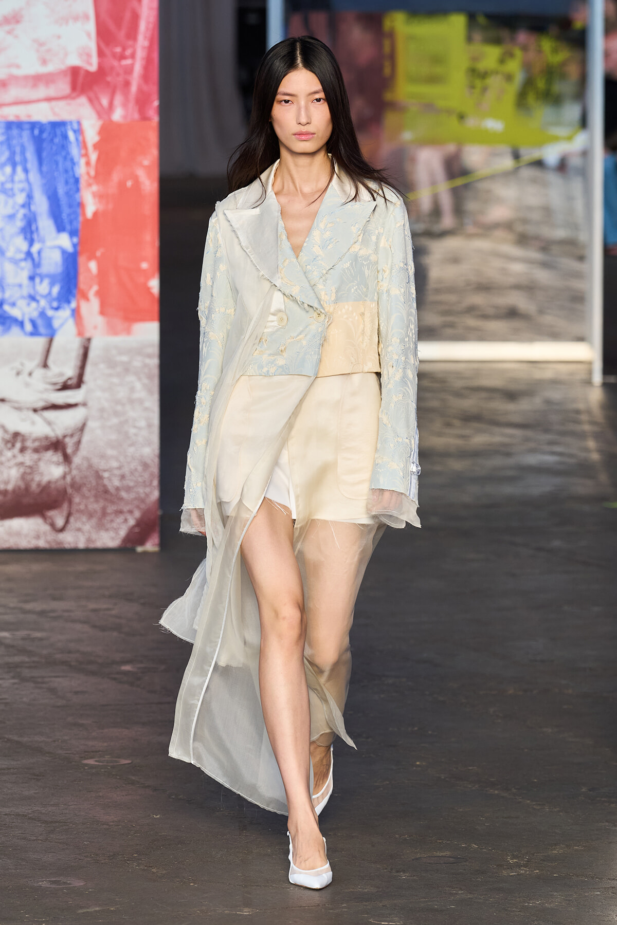 Female model walking a fashion runway in a pale blue-beige layered ensemble with a sheer cape and white heels.
