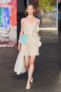 Model on runway wearing a beige strapless dress with patchwork panels and a blue fabric sash at the waist.