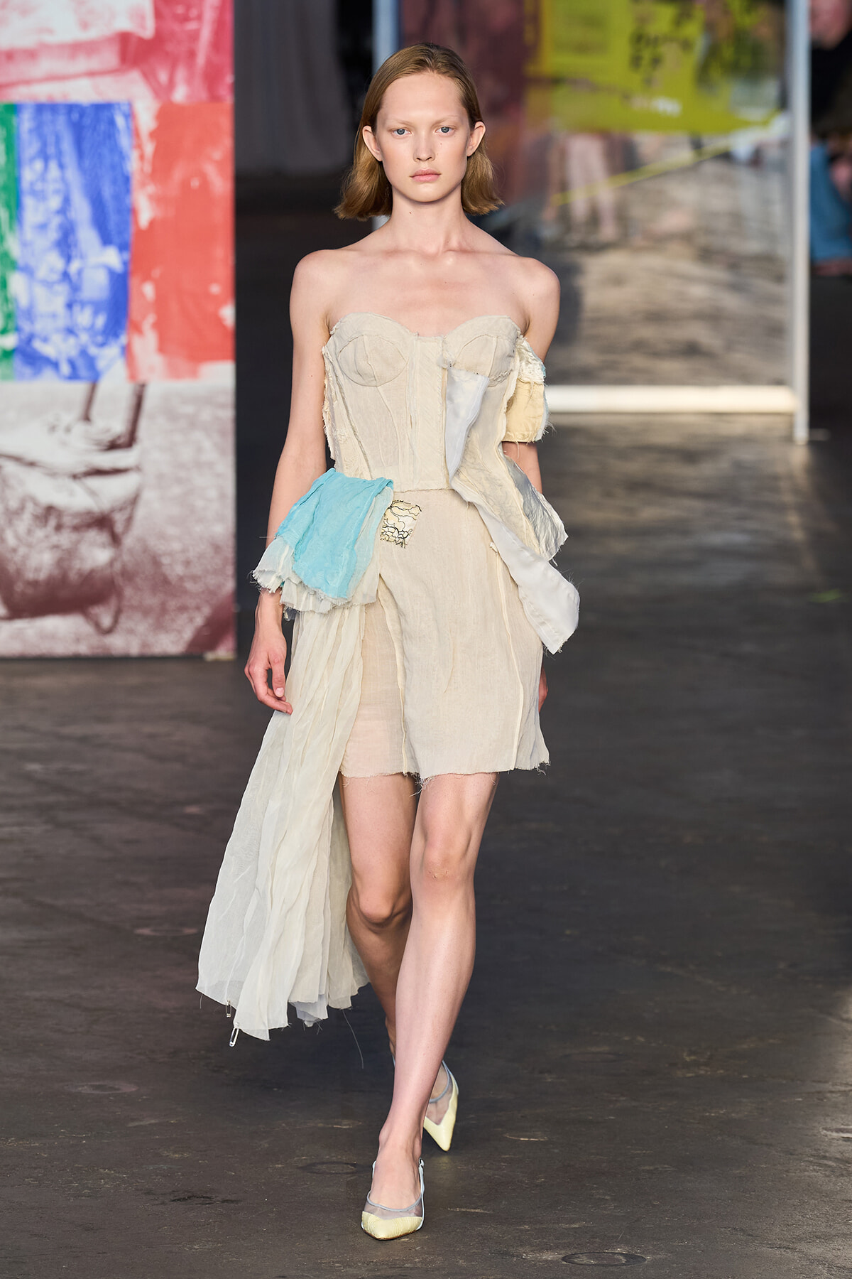 Model on runway wearing a beige strapless dress with patchwork panels and a blue fabric sash at the waist.