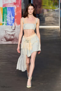 Model on a runway wearing a pale blue floral crop top and matching high-low skirt with a sheer draped panel.