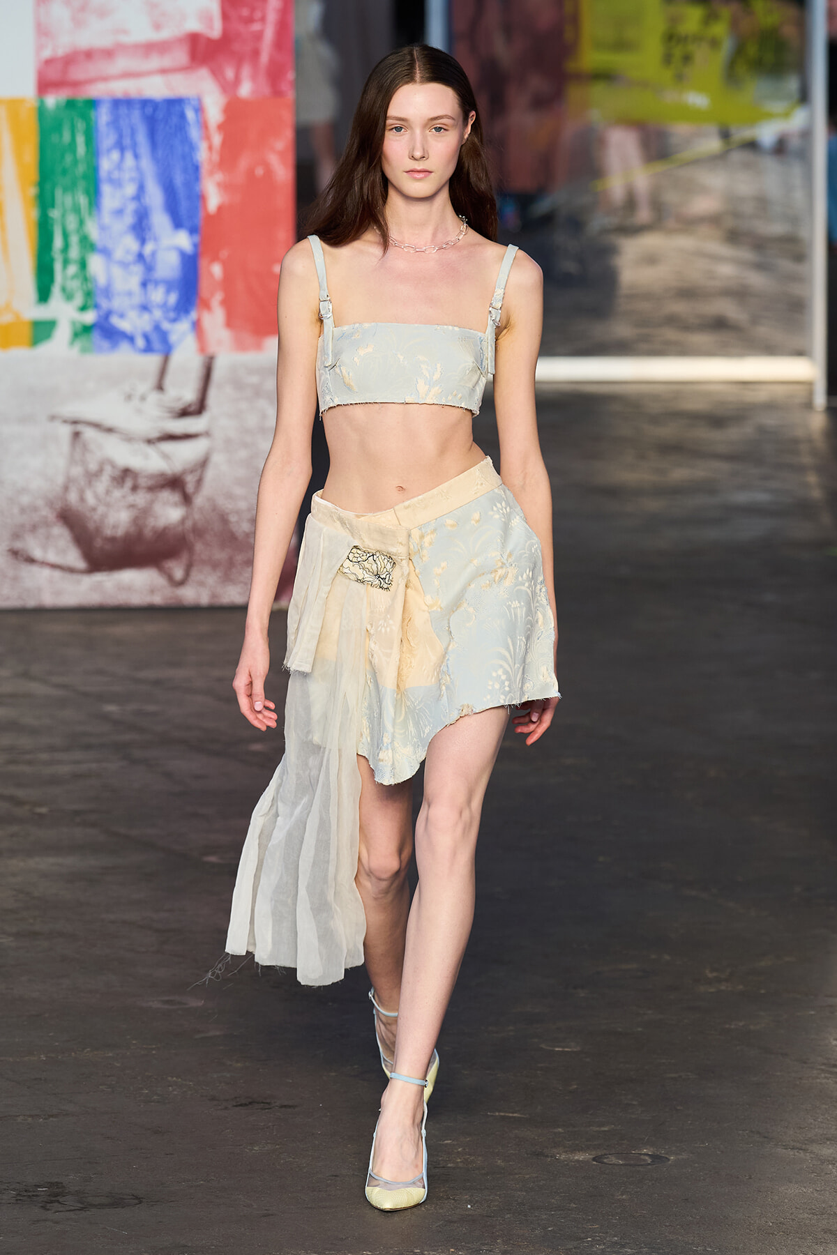Model on a runway wearing a pale blue floral crop top and matching high-low skirt with a sheer draped panel.