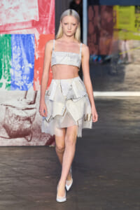 Model walking a runway in a white patchwork two-piece outfit: cropped top and asymmetrical layered skirt, wearing white heels, with blonde hair.