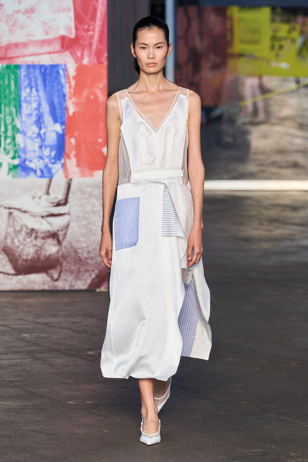 Model walking the runway in a white patchwork gown with sheer panels and blue striped pocket accents against a colorful backdrop.