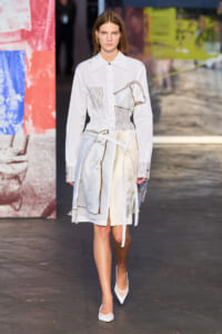 Model walks the runway in a white shirt-dress with patchwork panels and a waist belt, white heels, and a sculpted, abstract print.