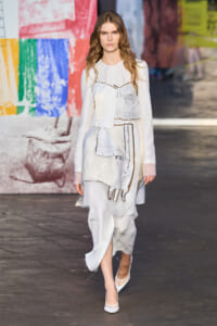 Model walking the runway in a white layered dress with patchwork panels and sheer sleeves, backdrop is a colorful mural graffiti wall.