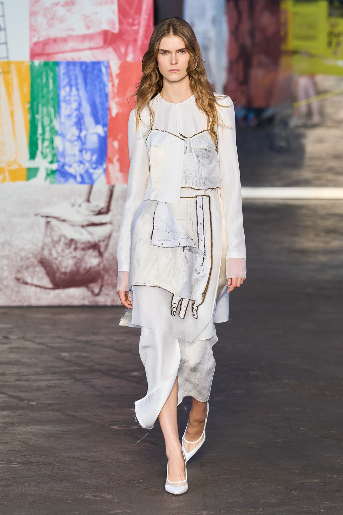 Model walking the runway in a white layered dress with patchwork panels and sheer sleeves, backdrop is a colorful mural graffiti wall.