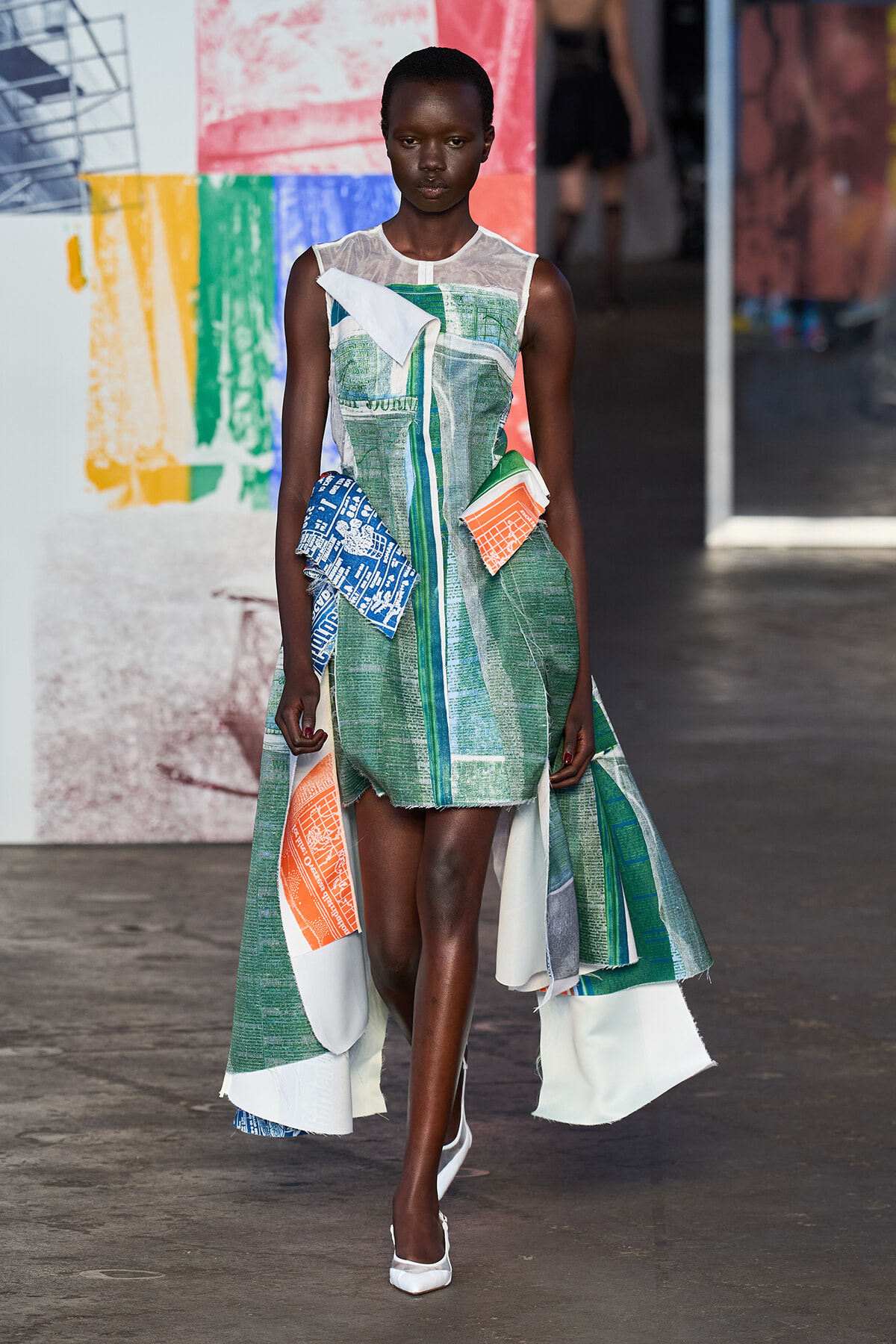 Model walks the runway in a green, white, and teal patchwork dress with orange and blue printed panels and white heels.