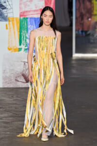 Model walking a fashion runway in a sheer sleeveless gown with vertical yellow, gold, and pale striped ribbons and a high slit.