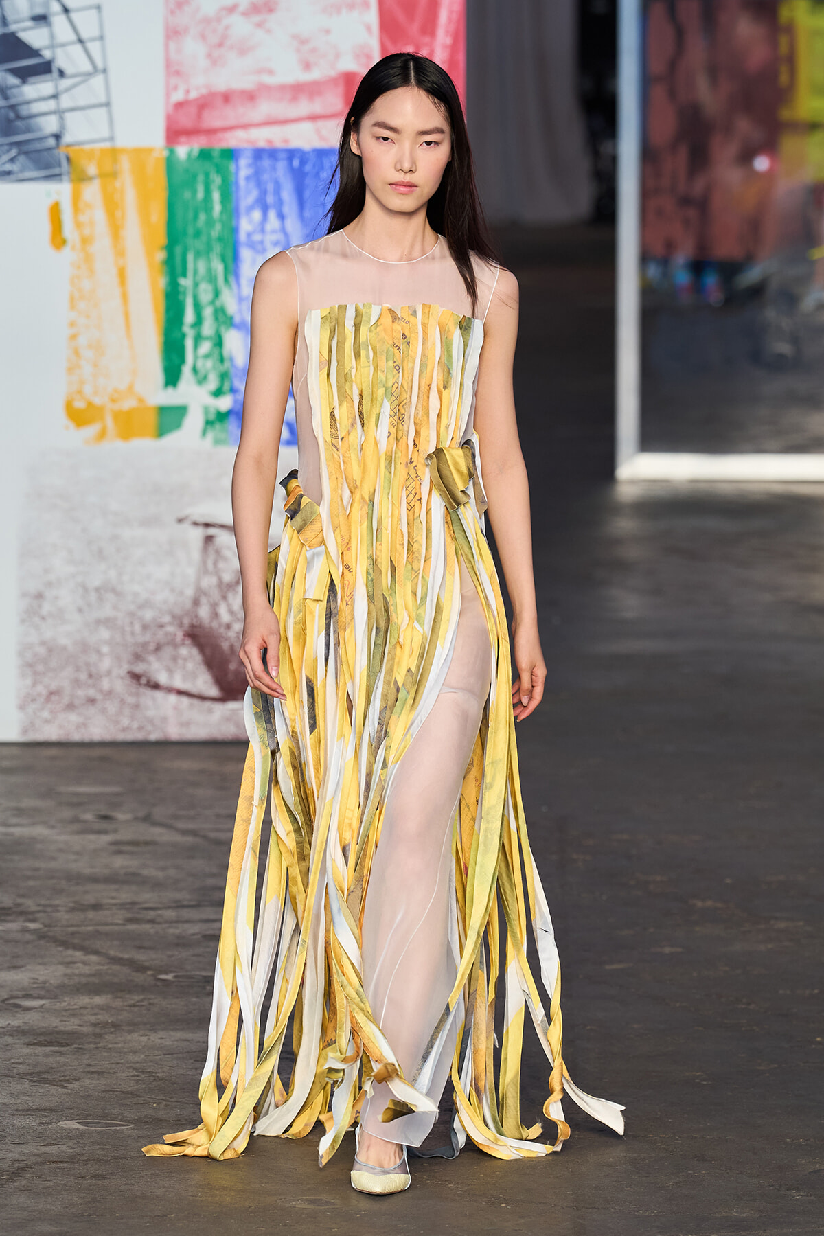 Model walking a fashion runway in a sheer sleeveless gown with vertical yellow, gold, and pale striped ribbons and a high slit.