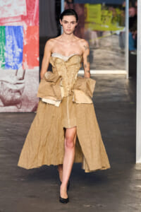 Model walks the runway in a beige strapless corset dress with layered fabric panels and a voluminous over-skirt, wearing black heels.