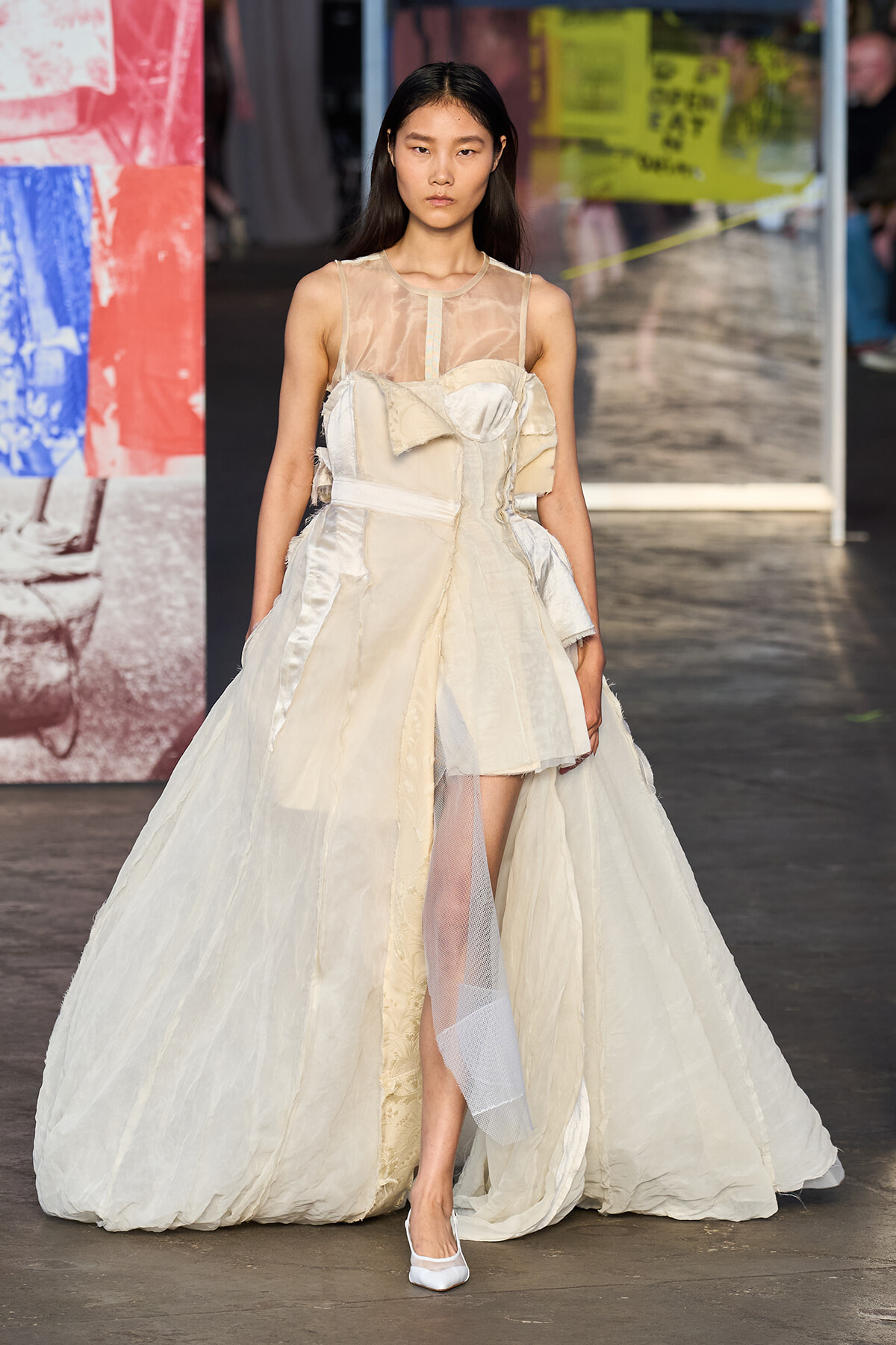 Fashion model on a runway wearing an ivory, layered ball gown with a sheer bodice and long train.