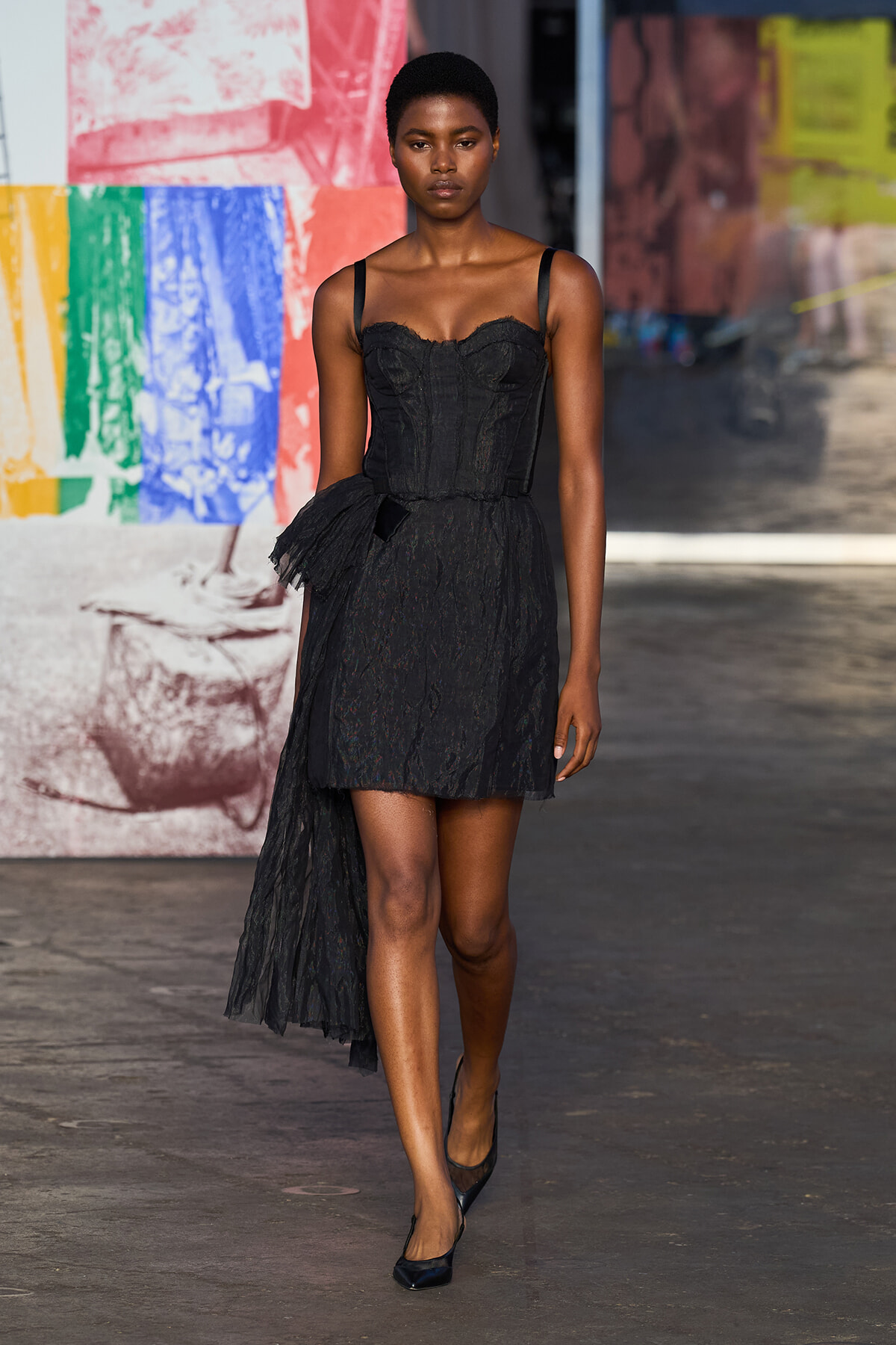 Model walking a runway in a black lace strap-shoulder dress with a short front and a sheer, longer train, against a colorful mural backdrop.