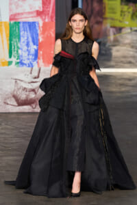 Model wearing a dramatic black ball gown with lace panels and a voluminous layered skirt on a fashion show runway onlookers blurred in the background