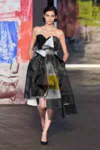 Model walks the runway in a strapless, patchwork black dress made of torn fabrics with white and colored panels, paired with black heels, against a colorful poster backdrop.