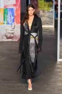 Runway model wears a long black patchwork coat with fringe panels and pale heels, posed before a colorful wall backdrop in a fashion show.