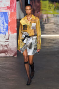 Model walks the runway in a yellow sheer patchwork blouse and newspaper-print wrap skirt, with a black belt and black heels.