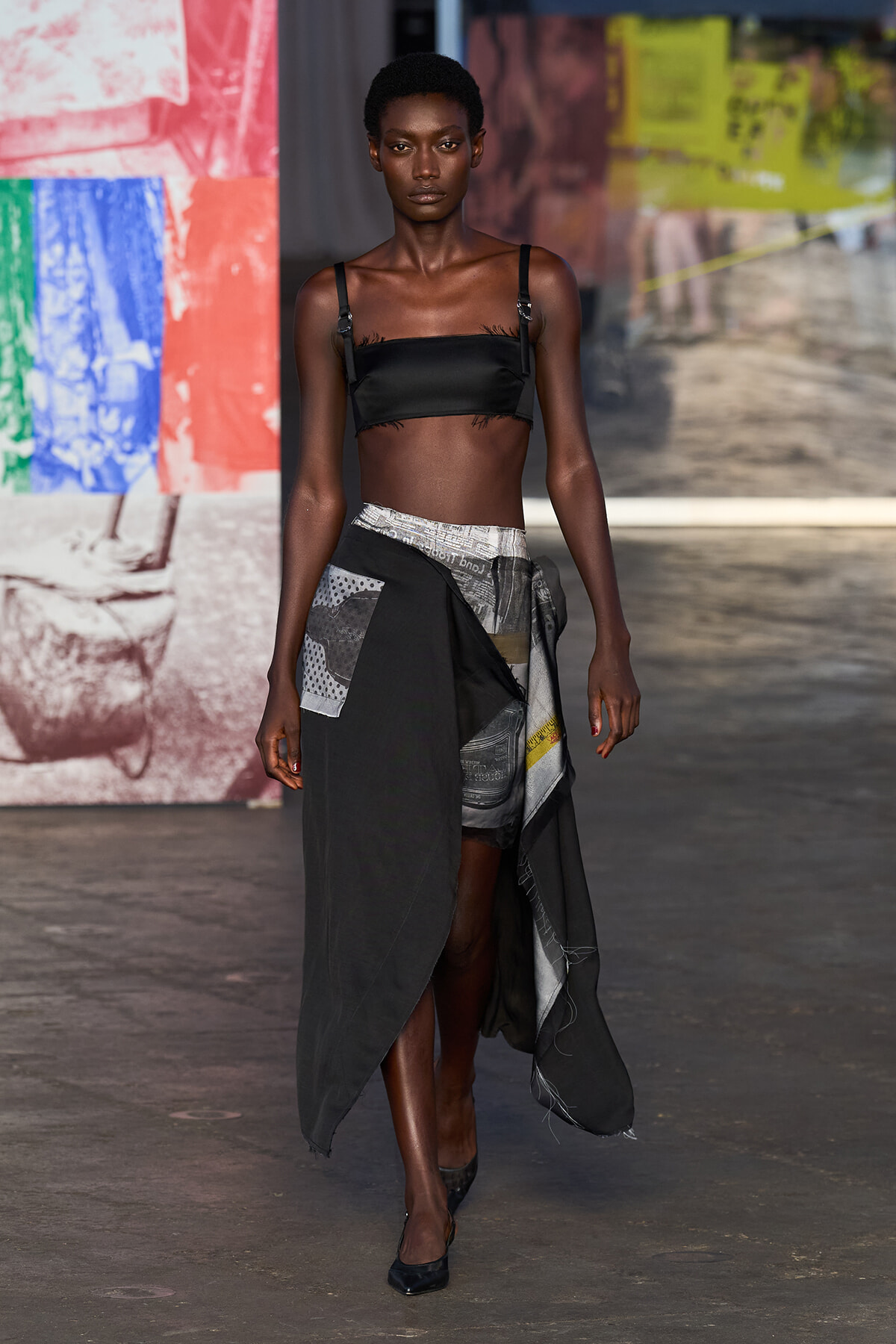 Model walks the runway in a black crop top with thin straps and an asymmetric, patchwork wrap skirt in grayscale tones.