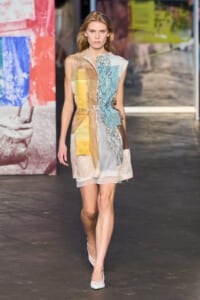 Model on a runway wearing a knee-length, color-block patchwork dress with lace panels and sheer hem, white heels.