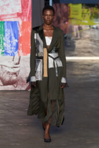 Female model walking runway in a long olive-green patchwork coat with a belt and layered fabrics, black heels.