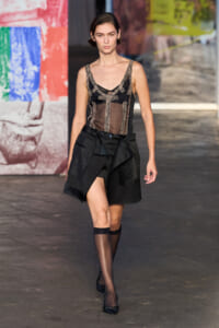 Model walks a fashion runway in a black sheer lace-trimmed top and an asymmetric black skirt with layered panels, black stockings and heels.