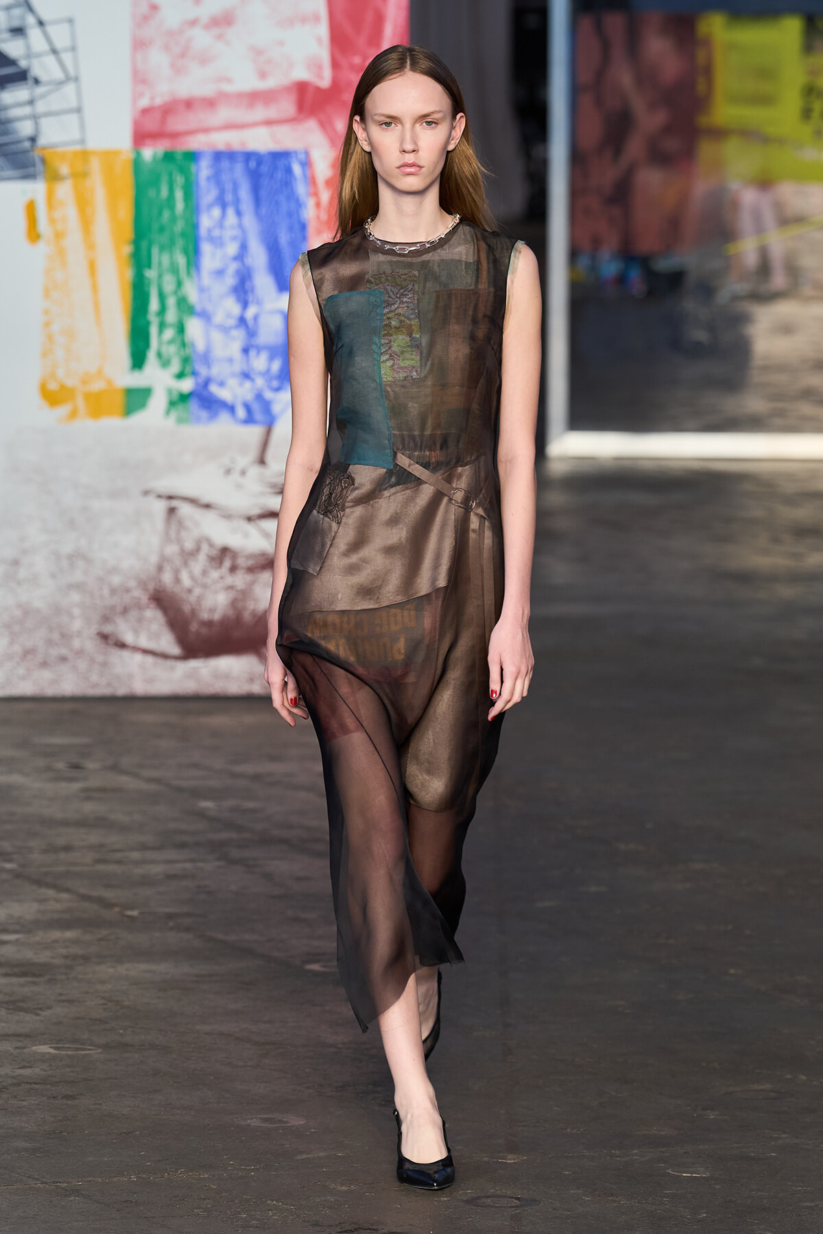 Female model walking a fashion show in a sheer, patchwork brown dress with black heels against a colorful backdrop.