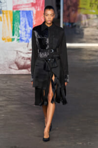Model on a fashion show runway wearing a black belted top with glossy panels and a fringed, uneven-hem skirt.