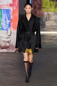 Model walks a fashion runway in a black belted coat over sheer yellow shorts, wearing black stockings and heels. Alternative: Model on a runway wearing an oversized black coat with a waist belt, paired with sheer yellow underlayers and black hosiery with heels. Alternative: Female model in a dark, structured coat belted at the waist, visible yellow sheer layer beneath, and black sheer socks with black heels. Alternative: Runway look featuring a black, cinched coat layered over a translucent yellow skirt short, with black tights and pumps. Alternative: Model in a monochrome black ensemble with a belted coat, peeking sheer yellow fabric, and black footwear.