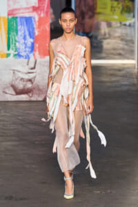 Model walking the runway in a sheer pastel dress with cascading multicolored ribbon strips down the front (fashion show).