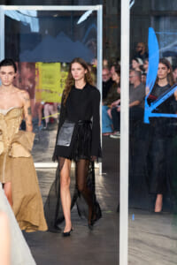 Model walks a runway wearing a black blazer and sheer black skirt as onlookers sit behind glass panels in the background, left model in a beige gown nearby.